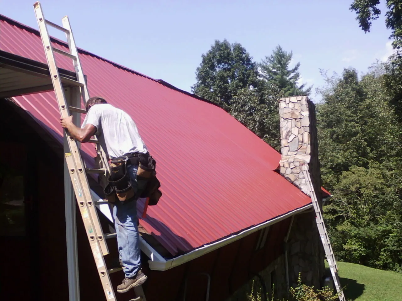 Roofer on a ladder beside a red metal roof and stone chimney — Soffit & Fascia Repair by Roofing Brothers in Presto